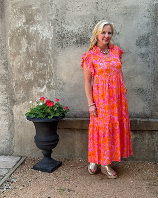 Woman in a pink and orange floral dress standing next to a stone wall with a pot of flowers.
