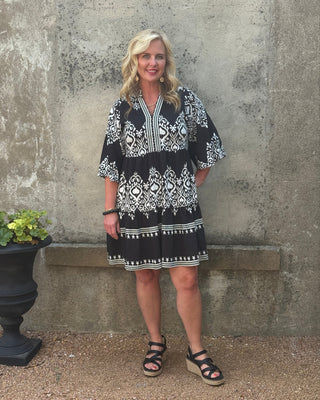 Woman wearing a black and white patterned dress standing against a textured wall.