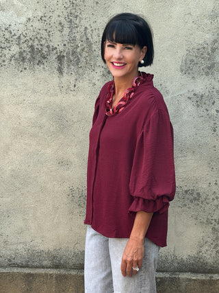 Woman wearing a burgundy blouse with ruffled sleeves against a textured wall.