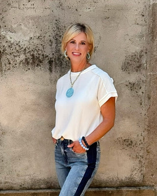 Woman in a white shirt and blue jeans standing next to a stone wall with a flower pot.