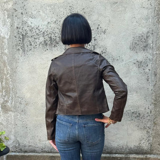 Person wearing a brown leather jacket and blue jeans standing against a textured wall.