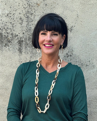 Woman wearing a green long-sleeve top and blue jeans with a long chain necklace against a textured wall.