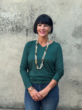 Woman wearing a green long-sleeve top and blue jeans with a long chain necklace against a textured wall.