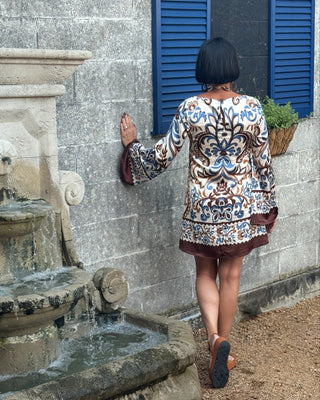 Woman in a patterned jacket standing next to a stone fountain against a gray wall with blue shutters.