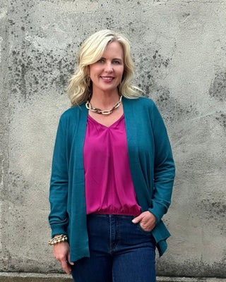 Woman wearing a blue cardigan, pink shirt, and dark jeans standing against a concrete wall.