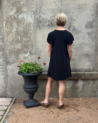 A woman wearing a black short-sleeve dress stands outdoors next to a concrete wall and a potted plant, viewed from above at an angle.
