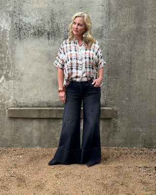 Woman wearing a plaid shirt and black flared pants standing against a textured wall.