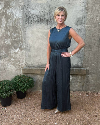 Woman in a dark blue dress standing against a textured wall with plants.