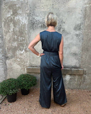 Person wearing a dark blue jumpsuit standing against a textured wall with potted plants.