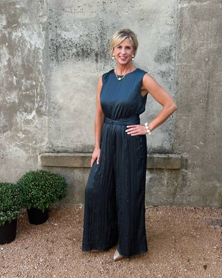 Woman in a dark blue sleeveless dress standing against a textured wall with plants.