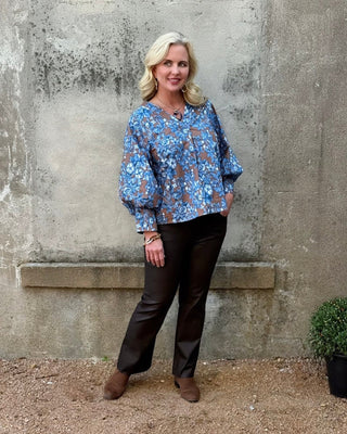 Woman wearing a blue floral blouse and dark pants standing against a textured wall.