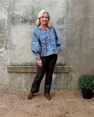 Woman wearing a blue floral blouse and dark pants standing against a textured wall.