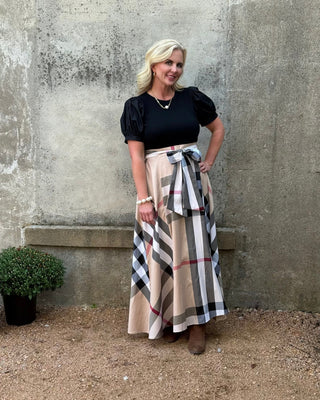 Woman wearing a black top and plaid skirt standing against a textured wall.