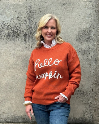 Woman wearing an orange sweater with 'hello my name is' text, standing against a concrete wall.