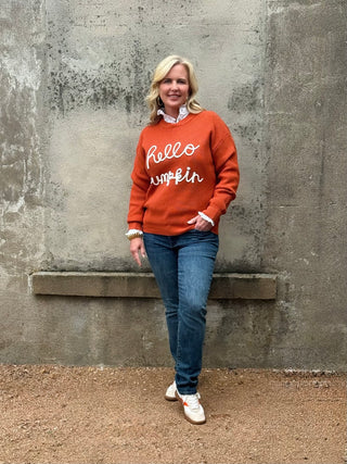 Woman wearing an orange sweater with 'Hello Pumpkin' text against a textured wall.