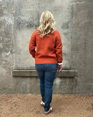 Person wearing an orange sweater and blue jeans standing against a gray wall.