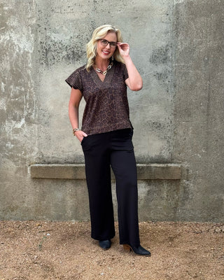 Woman wearing a patterned top and black pants against a textured wall.