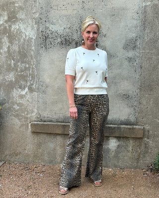 Woman wearing a white top and leopard print pants standing against a concrete wall.