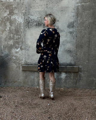 Person wearing a patterned dress and silver boots standing against a textured wall.