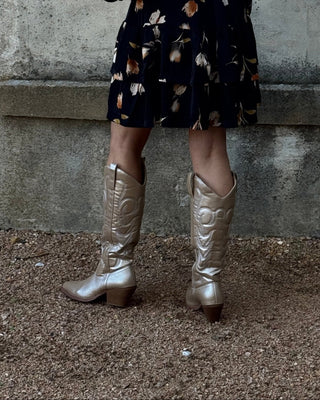 Woman in a patterned dress and metallic boots standing against a textured wall.