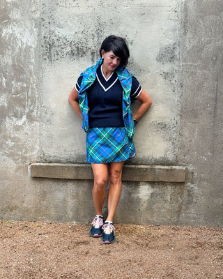 Person wearing a blue plaid skirt and vest against a concrete wall.