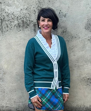 Woman in a green cardigan and plaid skirt standing against a concrete wall.