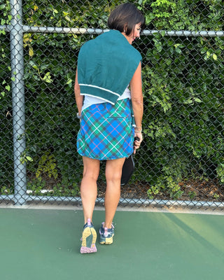 Person on a tennis court wearing a green towel over their shoulder and a blue plaid skirt.