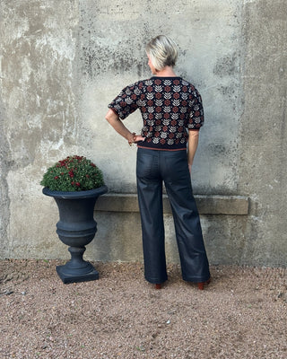 Person wearing a patterned top and dark pants standing against a textured wall with a planter nearby.