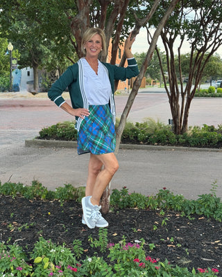 Woman in a plaid skirt and green cardigan standing outdoors near trees and flowers.