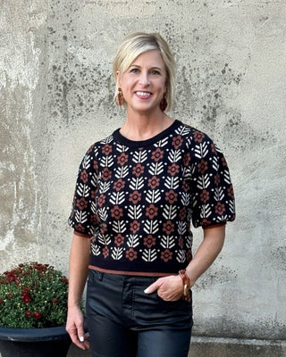 Woman wearing a patterned sweater and dark jeans standing in front of a textured wall with a plant.