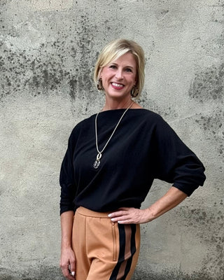 Woman wearing a black top and tan pants with black stripes standing against a textured wall.