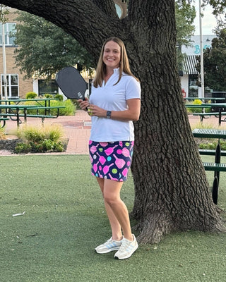 Woman holding a pickleball paddle standing next to a tree on a pickleball court.