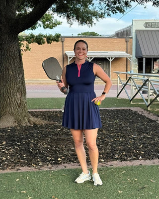 Woman in a navy dress holding a paddle on a pickleball court with a building and tree in the background.