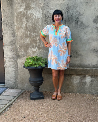 A woman stands outdoors against a textured concrete wall next to a black planter, wearing a light blue short-sleeve dress with orange and green floral patterns and a yellow-orange collar.