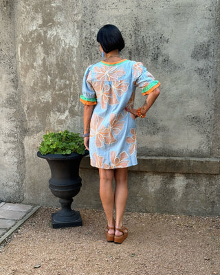 A woman stands with her back to the camera wearing a light blue short-sleeve dress with a peach and orange floral pattern and colorful trim. She is standing outdoors in front of a textured concrete wall next to a black planter pot.
