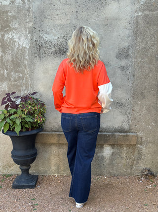 Person wearing an orange sweatshirt and blue jeans standing on a concrete surface with a plant nearby.