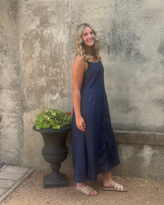 A woman wearing a sleeveless navy blue maxi dress and beige flat sandals, standing outdoors against a textured concrete wall.