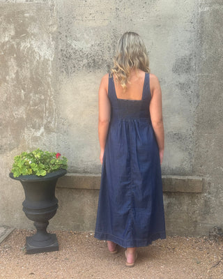 A woman wearing a sleeveless navy blue midi dress, standing outdoors next to a concrete wall and a black planter.