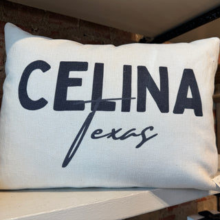 A lumbar style white canvas throw pillow with 'CELINA Texas' printed in dark navy lettering, resting on a white shelf against a brick wall background.