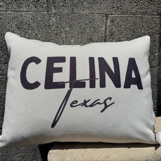 A lumbar style throw pillow with 'CELINA Texas' printed in black, resting on a stone ledge against a dark textured brick wall.