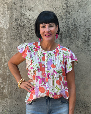 A woman with a black bob haircut is wearing a white short-sleeve blouse with a colorful floral print of orange, pink, and green, paired with light blue jeans, standing against a textured concrete wall.