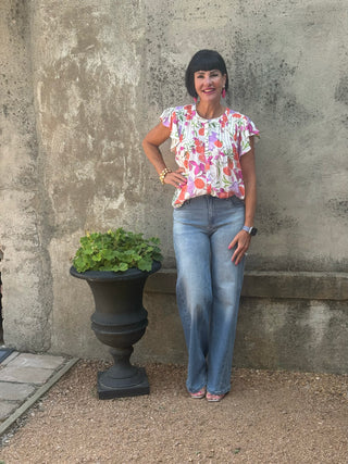 A woman with a black bob haircut stands outdoors against a textured concrete wall next to a large black planter with green leaves. She is wearing a colorful floral short-sleeve top and light blue jeans.