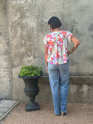 A woman viewed from behind wearing a short-sleeve floral print top and light blue jeans, standing in front of a textured concrete wall next to a black planter.