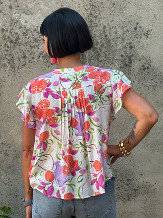 A woman viewed from behind wearing a short-sleeve floral blouse with a cream base and orange, pink, and purple flowers with green leaves, paired with blue jeans, standing against a textured concrete wall.