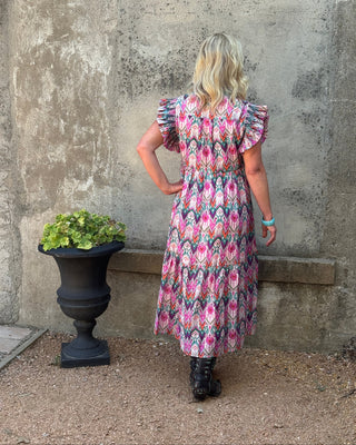 A woman stands with her back to the camera wearing a pink, blue, and white patterned midi dress with ruffled short sleeves, posing in front of a textured concrete wall next to a black planter.