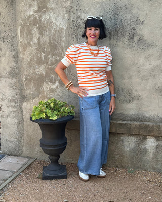 A woman with a black bob haircut and sunglasses on her head stands against a textured concrete wall wearing a white t-shirt with orange horizontal stripes, wide-leg blue jeans, and white sneakers. She is standing next to a black planter with green leaves.