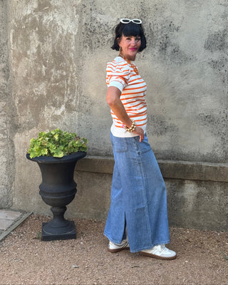 A woman posing outdoors against a textured concrete wall, wearing a white t-shirt with orange stripes, a long blue denim skirt, and white sneakers.