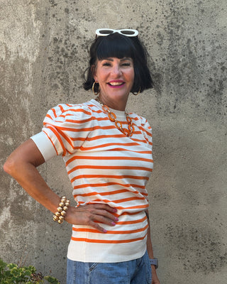 A woman with short black hair and white-framed sunglasses on her head, wearing a short-sleeve knit top with orange and white horizontal stripes, blue jeans, and gold jewelry, posing against a textured concrete wall.