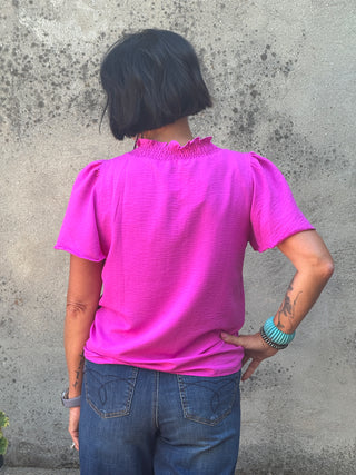 A woman viewed from behind wearing a bright pink short-sleeve blouse with puff sleeves and a smocked neckline, paired with blue jeans, standing against a textured concrete wall.