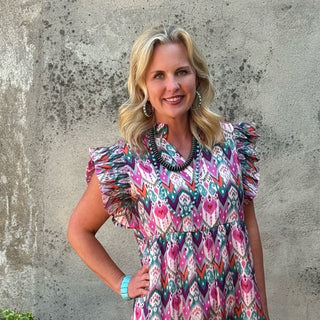 A woman stands outdoors against a textured concrete wall wearing a short-sleeved midi dress with a vibrant pink, purple, and blue floral pattern. She is wearing black ankle boots and standing next to a black planter with green leaves.